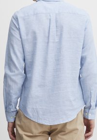 Vaaleansininen puuvillapaita pitkähihaisena, jossa on selässä laskostus, pyöristetty helma ja napilliset hihansuut. Teksturoitu kudonta tuo hienoista syvyyttä.