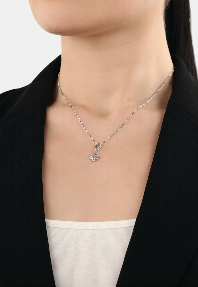 Collana in argento con un design floreale, impreziosita da piccoli cristalli, su una delicata catena, indossata sopra una maglietta bianca e un blazer nero.