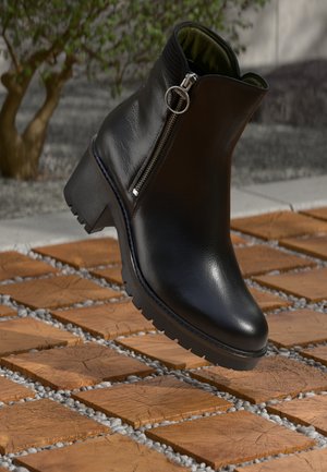 Černá kožená kotníková bota s bočním zipem, texturovaným svrškem, masivním podpatkem a robustní podrážkou. Obklopena vzorovanou dlaždicovou plochou.