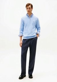 Νεαρός άνδρας φορά ανοιχτό μπλε πουλόβερ με φερμουάρ μέχρι το στήθος πάνω από ριγωτό πουκάμισο, σκούρες ναυτικές παντελόνες και μαύρα παπούτσια, στέκεται με το ένα χέρι στην τσέπη.