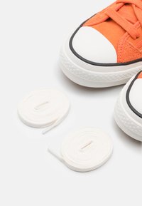 Pomarańczowe zamszowe trampki z białymi gumowymi podeszwami i czarnymi akcentami. Obok butów znajdują się dwa białe sznurówki.