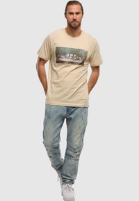 Camiseta de algodón beige con un gráfico de "La Última Cena", combinada con jeans de denim azul claro desgastados y zapatillas blancas.