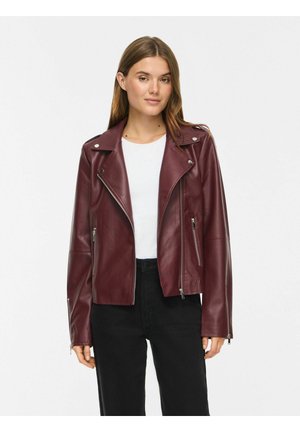 Jeune femme portant une veste en cuir bordeaux par-dessus une chemise blanche et un pantalon noir, debout devant un fond blanc uni.