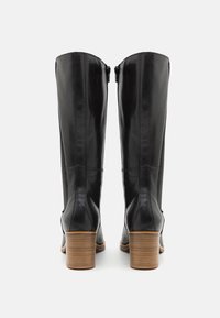 Botas altas de cuero negro con textura suave, cremalleras laterales y un tacón de madera apilada. La vista trasera resalta el diseño elegante.