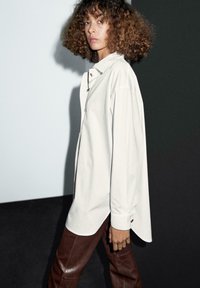 Camicia bianca oversize con bottoni dorati, abbinata a pantaloni di pelle marroni. Presenta maniche lunghe e una vestibilità comoda.