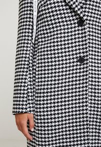 Czarnobiały płaszcz w wzór jodełki z czarnymi guzikami, noszony przez osobę z ręką spoczywającą obok płaszcza.