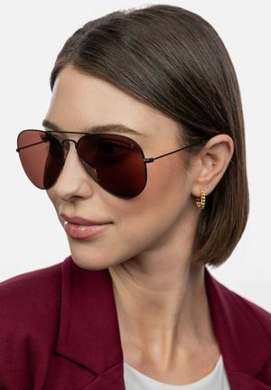 Mørke aviator-solbriller med brune linser og tynde sorte metalrammer. Modellen har en kort bob-hårfrisure og bærer en bordeaux blazer samt guldøreringe.