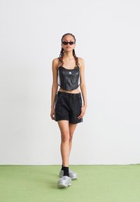 Top cropped de couro sintético preto com alças ajustáveis, combinado com calções de ganga pretos e ténis prateados. Modelo usa óculos de sol escuros.