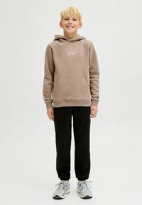Svijetlosmeđa hoodie s prednjim džepom i bijelim natpisom, uparen s crnim trenirkama i sivim tenisicama, stoji protiv jednostavne pozadine.