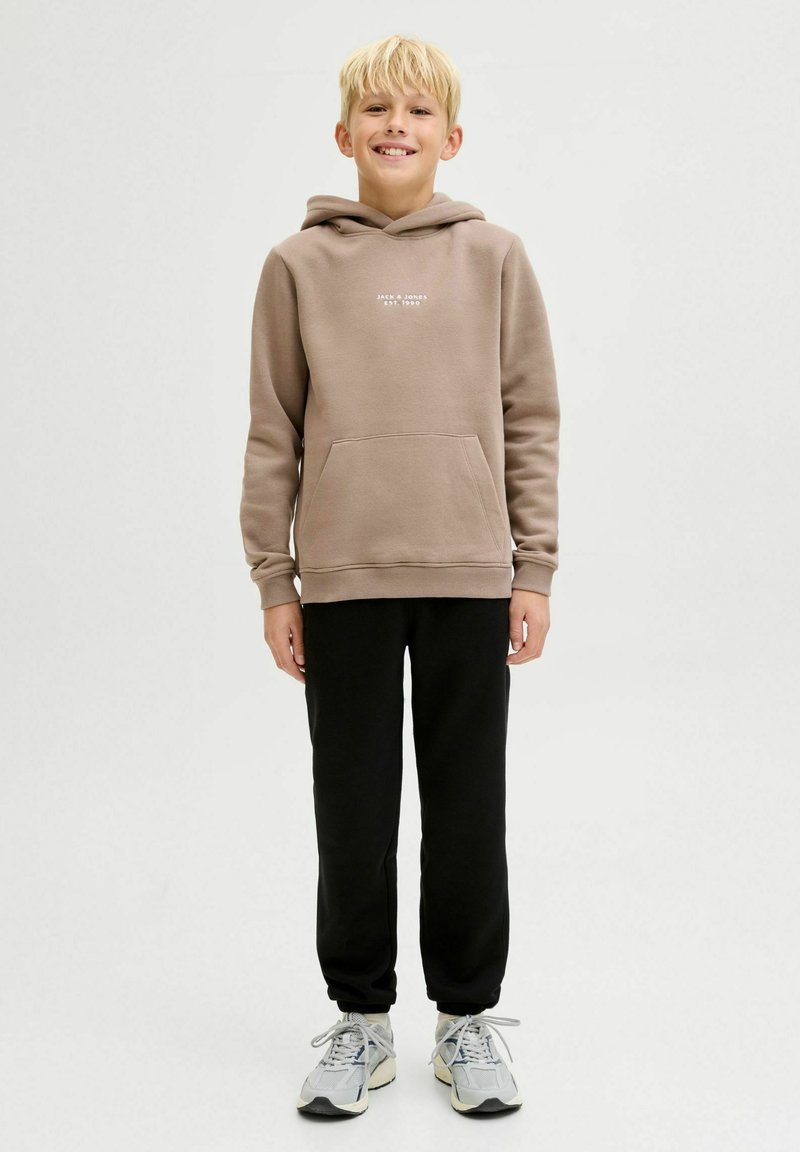 Svijetlosmeđa hoodie s prednjim džepom i bijelim natpisom, uparen s crnim trenirkama i sivim tenisicama, stoji protiv jednostavne pozadine.