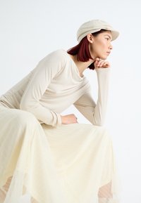 Langærmet, cremefarvet top med draperet halsudskæring, parret med en plisseret, gennemsigtig, lysegul nederdel. Modellen bærer en lys beige kasket.