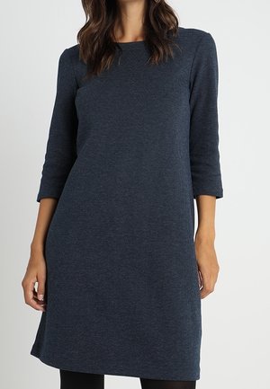 Mujer con un vestido gris oscuro simple hasta la rodilla con mangas tres cuartos sobre un fondo blanco liso.