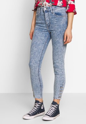 Femme portant un jean skinny bleu clair délavé à l'acide avec des boutons aux chevilles, un chemisier rouge à fleurs et des baskets montantes bleu marine.