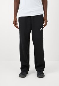 Mand iført sorte Adidas træningsbukser med hvidt tre-stribe logo og sidestropper, sorte sneakers og en hvid skjorte, stående på hvid baggrund.