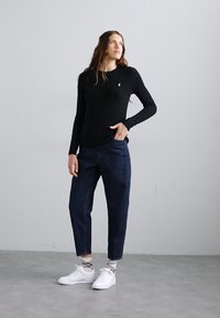 Črn pulover z pletenim vzorcem, mornarsko modre hlače, bele superge. Model stoji pokončno, hlače so sproščenega kroja, z diskretnimi teksturami in oblikovalskimi detajli.