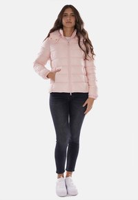 Giacca puffer rosa imbottita con zip frontale, tasche laterali e cappuccio staccabile, abbinata a jeans scuri e sneakers bianche.