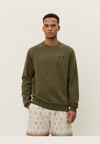 Olivgrüner Pullover aus weichem Material, mit rundem Halsausschnitt und einem kleinen schwarzen Logoetikett auf der Brust, kombiniert mit gemusterten Shorts.