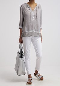 Blouse gris clair à col en V avec cordons, pantalon blanc, sandales argentées et sac à main gris texturé avec des accents noirs.