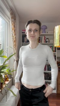 Személy áll bent, az ablak mellett, növényekkel, fehér hosszú ujjú ingben és fekete nadrágban, a háttérben könyvespolccal és lámpával.
