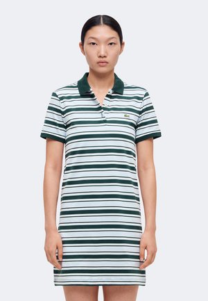 Jeune femme debout, tournée vers l'avant, portant une robe polo à manches courtes rayée horizontalement vert et blanc avec un col vert foncé.