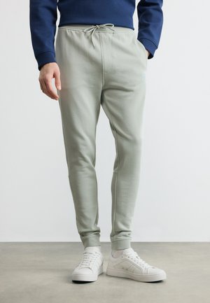 Man draagt lichtgrijze joggerbroek met trekkoord in de taille, gecombineerd met witte sneakers en een donkerblauw shirt met lange mouwen.