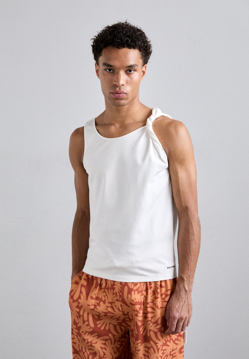 Top senza maniche bianco con un dettaglio di spalla intrecciato, abbinato a pantaloni corti arancioni con un motivo a corallo.