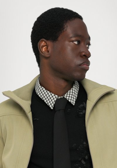 Olive green jacket with a zip and textured collar over a black sweater with distressing, layered with a white checkered shirt and black tie.