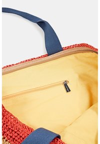 Intérieur d'un sac cabas tissé rouge avec doublure en tissu jaune, poche intérieure zippée et poignées bleu marine visibles.