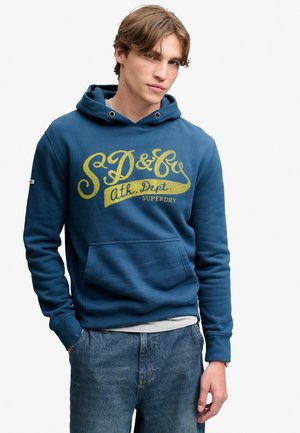 Jonge man draagt een blauwe Superdry capuchontrui met gele letters en een blauwe spijkerbroek, staand met één hand in de zak.
