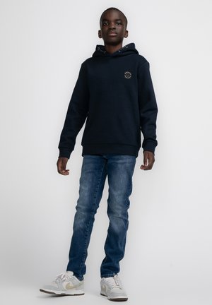 Marineblauwe hoodie van zachte stof, met een logo op de borst; gecombineerd met jeans van gemiddelde wassing en witte sneakers met grijze en gouden accenten.