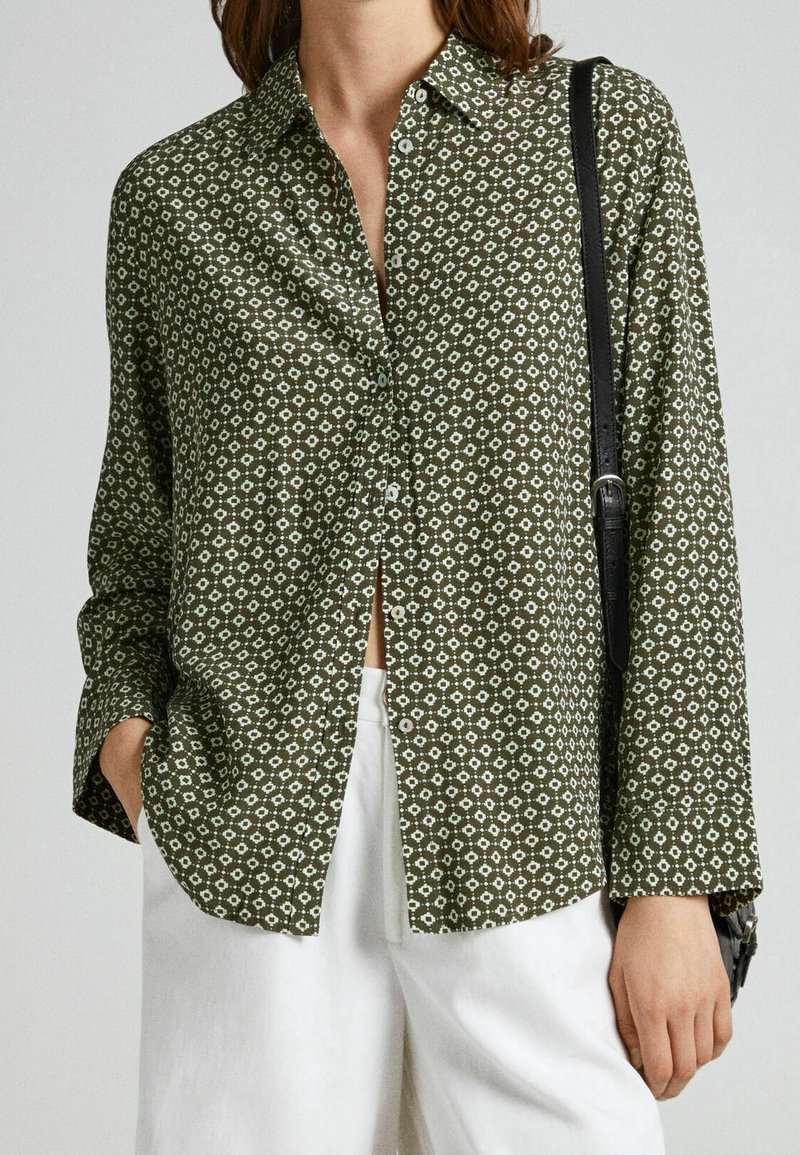 Personne portant une chemise boutonnée à motifs vert olive avec un pantalon blanc et un sac noir à l'épaule, main dans la poche.
