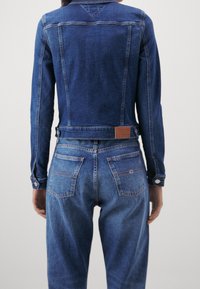 Modrá džínová bunda s přiléhavým střihem, s knoflíkovými manžetami a hnědou koženou etiketou na zadní straně. Spárována s odpovídajícími džínami s vysokým pasem.