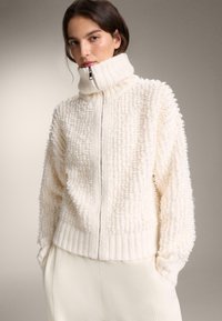 Krémová huňatá bunda na zips s vysokým rebrovaným golierom, textúrovanou látkou a rebrovaným lemom. Model ju nosí s nohavicami svetlej farby.