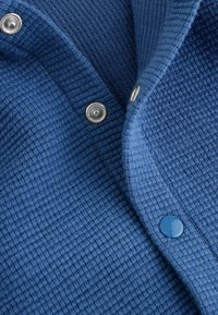 Close-up of blue textured fabric with metal and blue snap buttons fastened along the edge of a garment.