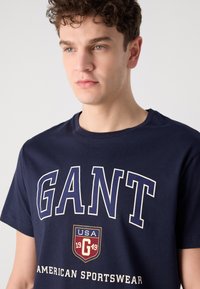 Tricou din bumbac bleu marin, cu un logo îndrăzneț "GANT" în alb, având un emblemă care afișează "USA 1949" dedesubt. Textură moale, design cu guler rotund.