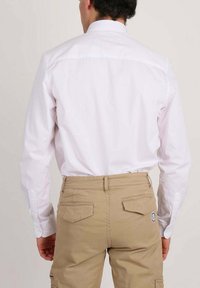 Un homme portant une chemise blanche à manches longues rentrée dans un pantalon beige avec des poches arrières à rabat et un petit patch rond sur la poche droite.