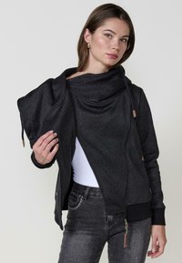 Giacca nera con texture e scollatura drappeggiata, dotata di chiusura con zip e coulisse, abbinata a jeans scuri e una maglietta bianca.