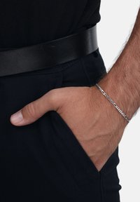 Bracciale in argento su un polso, appoggiato in una tasca. La mano è posizionata con il pollice visibile. La cintura in pelle nera contrasta con i pantaloni in tessuto blu navy.