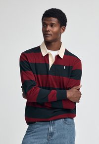 Rot gestreiftes Langarm-Polo-Shirt in Navy mit einem beigen Kragen und einem kleinen Logo auf der linken Brust. Weicher Stoff, entspannte Passform.