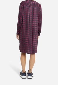 Sukienka z guzikami, uszyta z ciemnej tkaniny o geometrycznym wzorze w czerwone i różowe kolory, z długimi rękawami i bocznymi rozporkami. Noszona z trampkami.