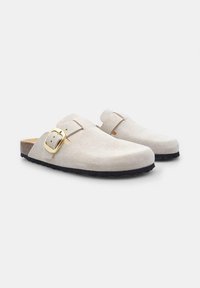 White suede clogs with rounded toes, a single buckle strap, brown cork footbed, and textured black rubber sole. Minimalistic design.