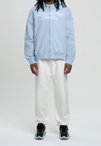 Sudadera con cremallera de color azul claro con texto bordado en blanco, combinada con pantalones de chándal crema y zapatillas deportivas negras con acentos blancos.