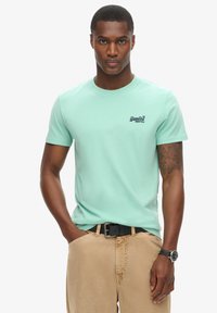 Mintgrøn bomulds-T-shirt med rund halsudskæring og korte ærmer, prydet med et lille mørkt logo på brystet, båret med beige bukser og et bælte.