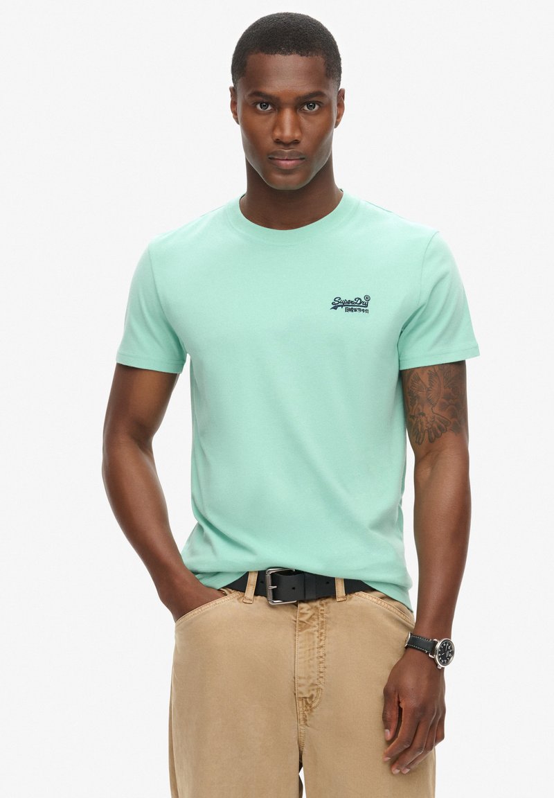 Mintgrøn bomulds-T-shirt med rund halsudskæring og korte ærmer, prydet med et lille mørkt logo på brystet, båret med beige bukser og et bælte.