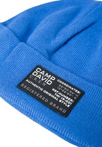 Blue knit beanie with a textured finish. Features a black rectangular label with white text and coordinate details, adding to the design.