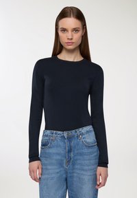 Długi rękaw, granatowy sweter z okrągłym dekoltem, w zestawie z jasnoniebieskimi spodniami typu high-waist, które mają klasyczne zapięcie na guziki i zamek błyskawiczny.