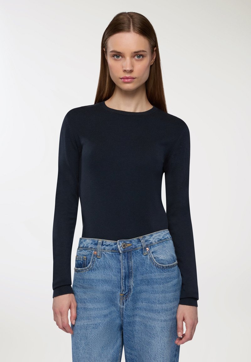 Długi rękaw, granatowy sweter z okrągłym dekoltem, w zestawie z jasnoniebieskimi spodniami typu high-waist, które mają klasyczne zapięcie na guziki i zamek błyskawiczny.