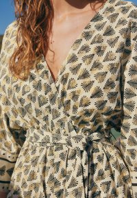 Robe portefeuille en tissu crème, ornée de motifs de feuilles noires et dorées, décolleté en V, manches courtes et taille nouée. Tissu texturé.