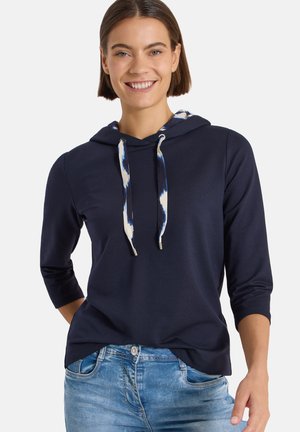 Donna sorridente che indossa una felpa con cappuccio blu navy con coulisse fantasia e jeans blu, in piedi con una mano in tasca su sfondo bianco.