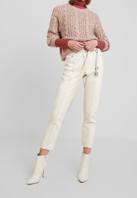 Personne portant un col roulé rose sous un pull rose texturé, un pantalon crème taille haute avec une longue ceinture, et des bottines à talons crème.